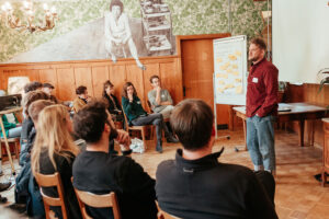 Tobias Bockstaller steht vor einer Runde Workshopteilnehmenden in einem Holzgetäferten Saal neben einem Flip-Chart voller Post-its.