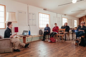In einem länglichen Saal mit weissen Wänden erläutert Christine Mühlebach etwas. Ihr gegenüber sitzen Workshopteilnehmende in looser Ordnung. An den Wänden hängen Flip-Chart-Bögen